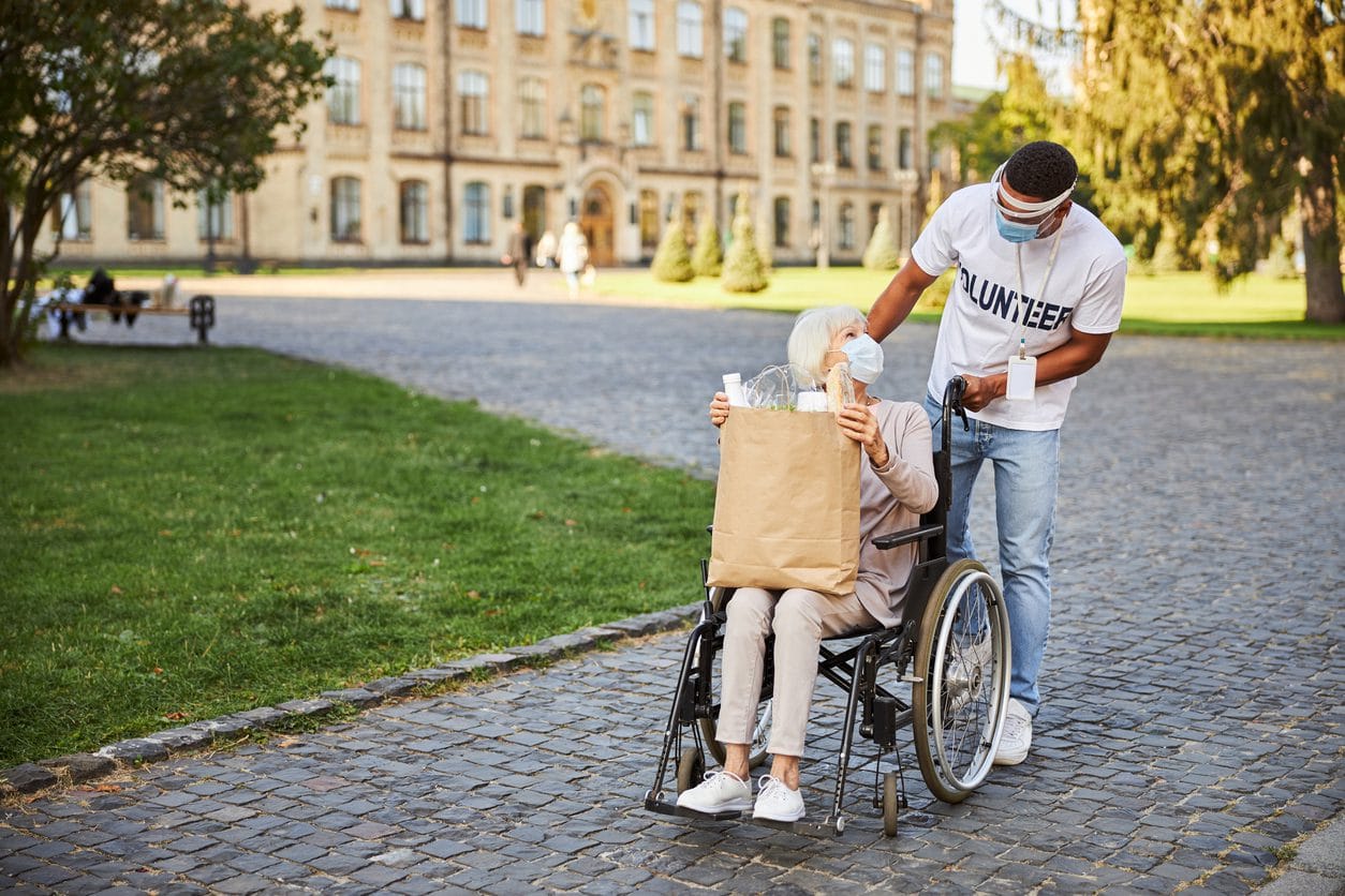 Reliable volunteer wearing a protective shield while leaning towards the elderly lady in wheelchair with a paper bag after shopping